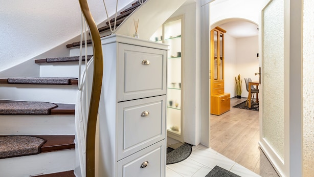Un escalier avec des marches en bois et un mur blanc, menant à une pièce où se trouvent un meuble en bois et une vitrine.