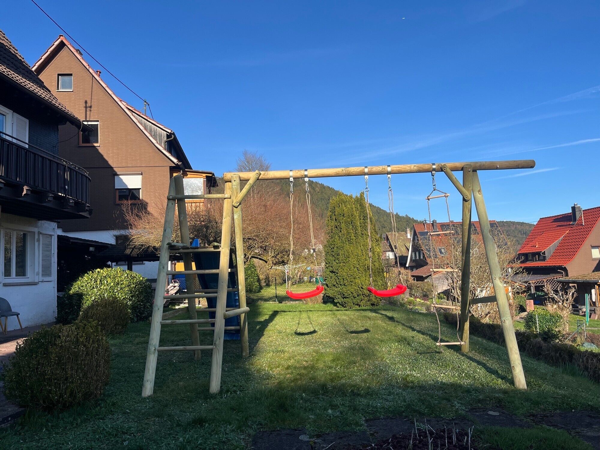 Ein Holzschaukelspielplatz in einem grünen Garten mit Häusern im Hintergrund.