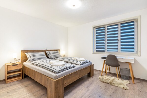 Une chambre à coucher avec un lit en bois, un bureau, une chaise, une table de chevet et une fenêtre avec des stores.