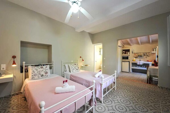 A room with two beds, a ceiling fan, a small table with a lamp, and a kitchenette in the background. - Stefanos Cottage in Paxi (Paxos)