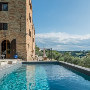A pool area with a clear blue pool, a brick building with arched doorways, and a view of rolling hills in the background. - Casatorre 6 2 Emma Villas (Saludecio)