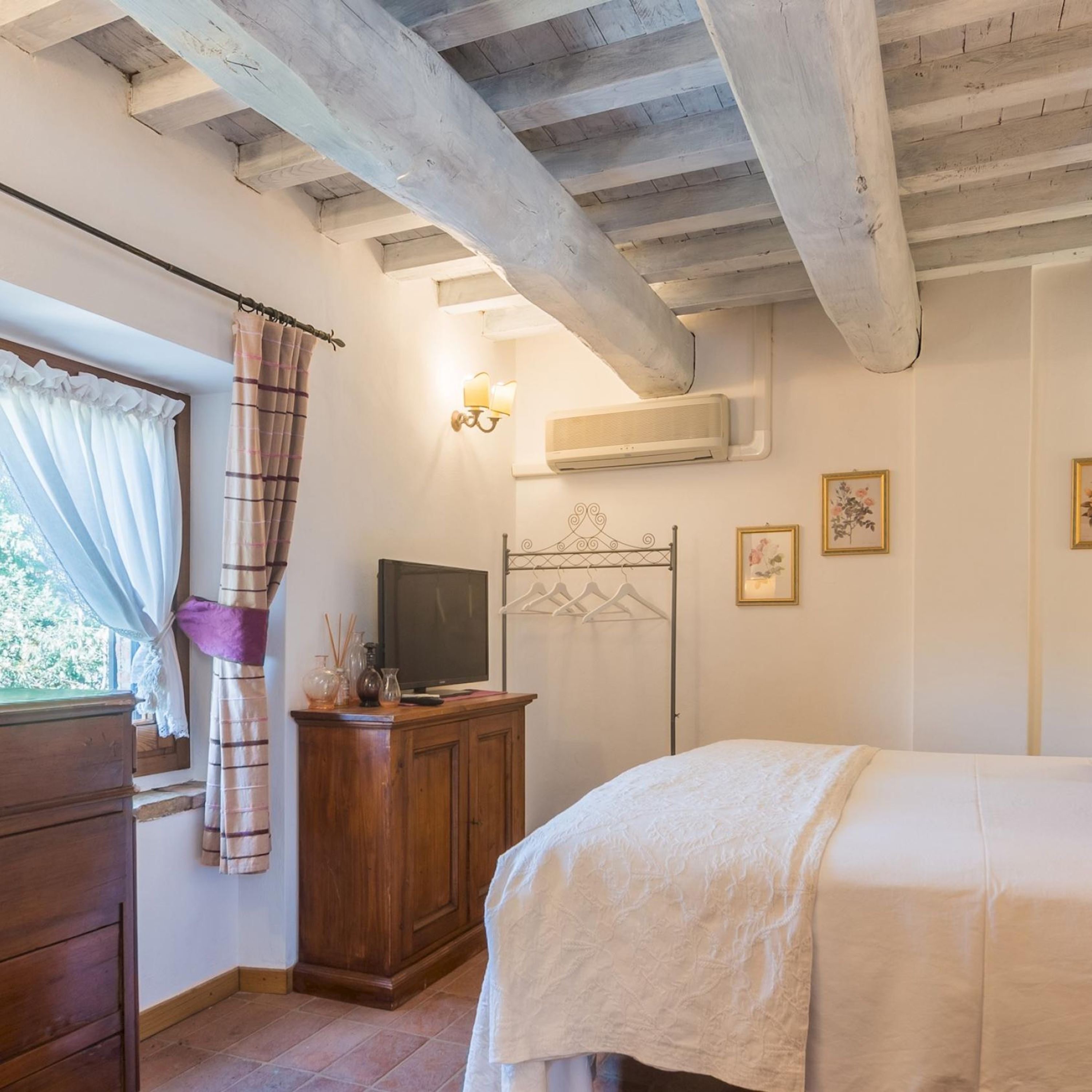 A bedroom with a wooden ceiling, a bed, a TV on a wooden cabinet, a window with curtains, and a wall-mounted air conditioner.