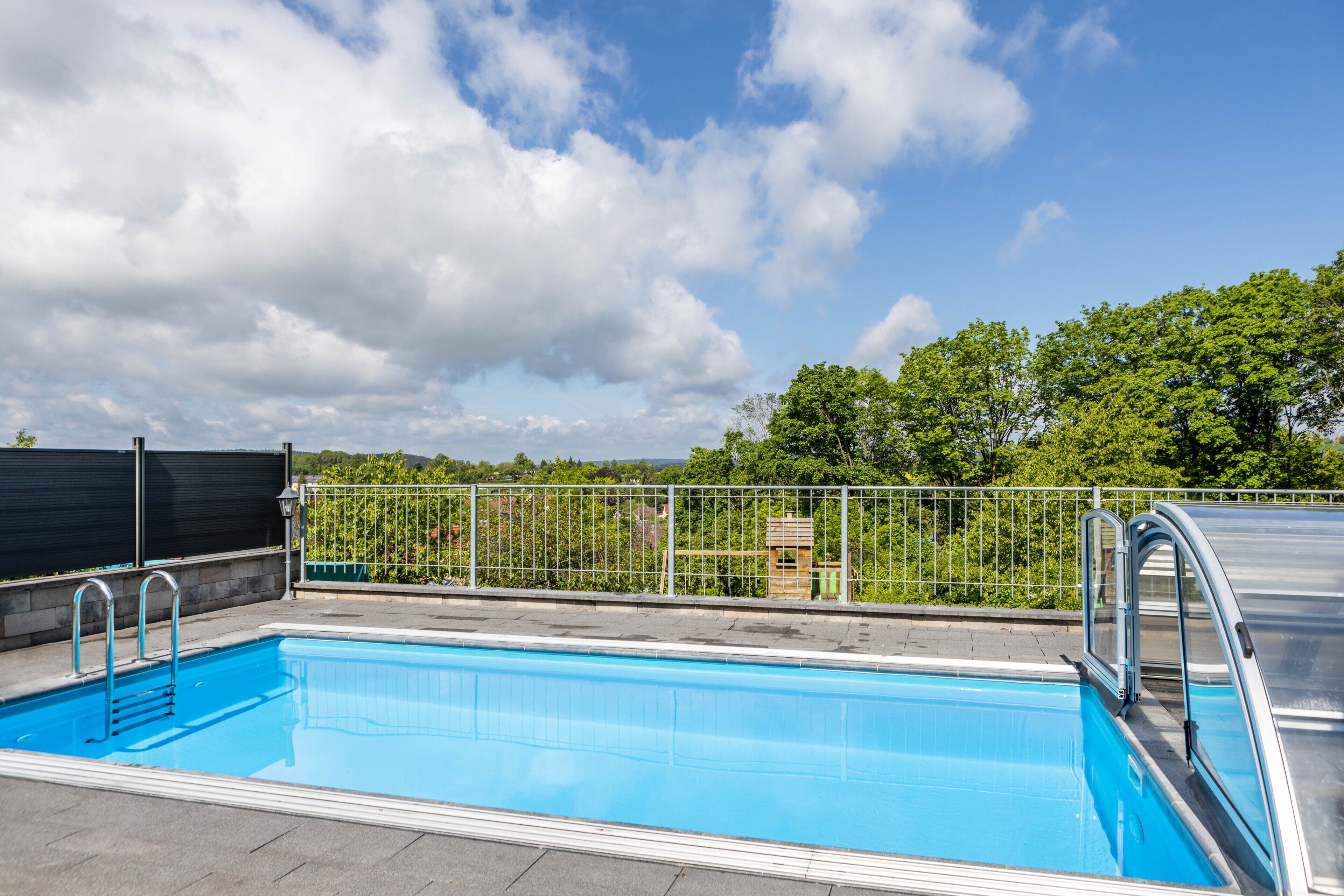 Une piscine entourée d’une clôture métallique, avec un espace abrité et une vue sur les arbres et les bâtiments en arrière-plan.