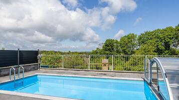Une piscine entourée d’une clôture métallique, avec un espace abrité et une vue sur les arbres et les bâtiments en arrière-plan.