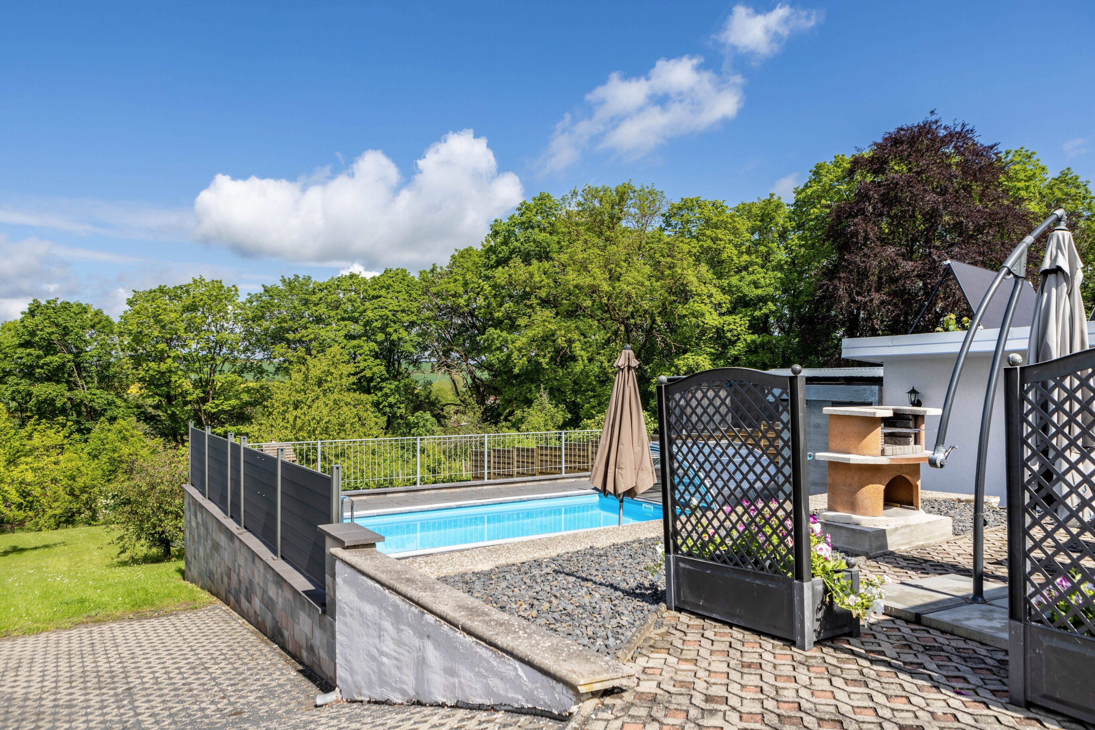 Une piscine entourée d’une clôture, un espace patio avec un barbecue et une vue sur les arbres.