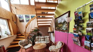 A room with a wooden staircase, wicker furniture, a pink wall with postcards, and a window with plants.