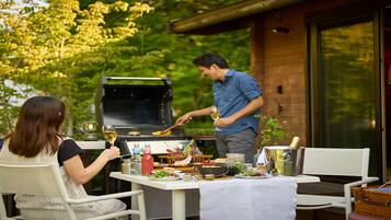 Un espacio de comedor al aire libre con una mesa dispuesta para dos personas, una parrilla y vistas a un edificio de madera.
