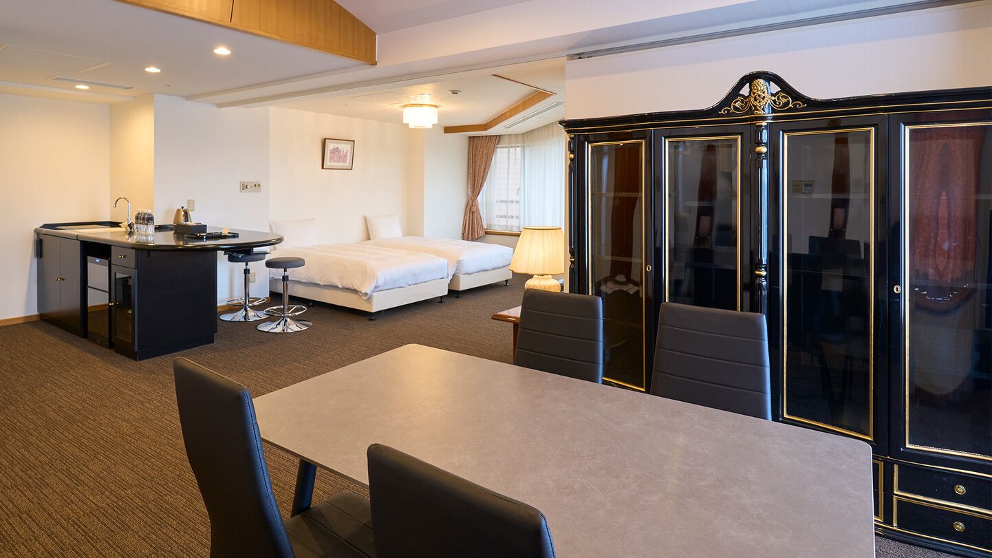 A hotel room with a dining table, two beds, a wardrobe, and a small kitchenette.