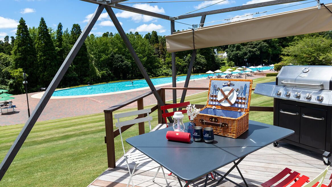 A deck with a grill, a table with a basket and bottles, and a view of a pool.
