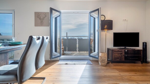A modern living room with a large window, a flat-screen TV, and a dining area with chairs.