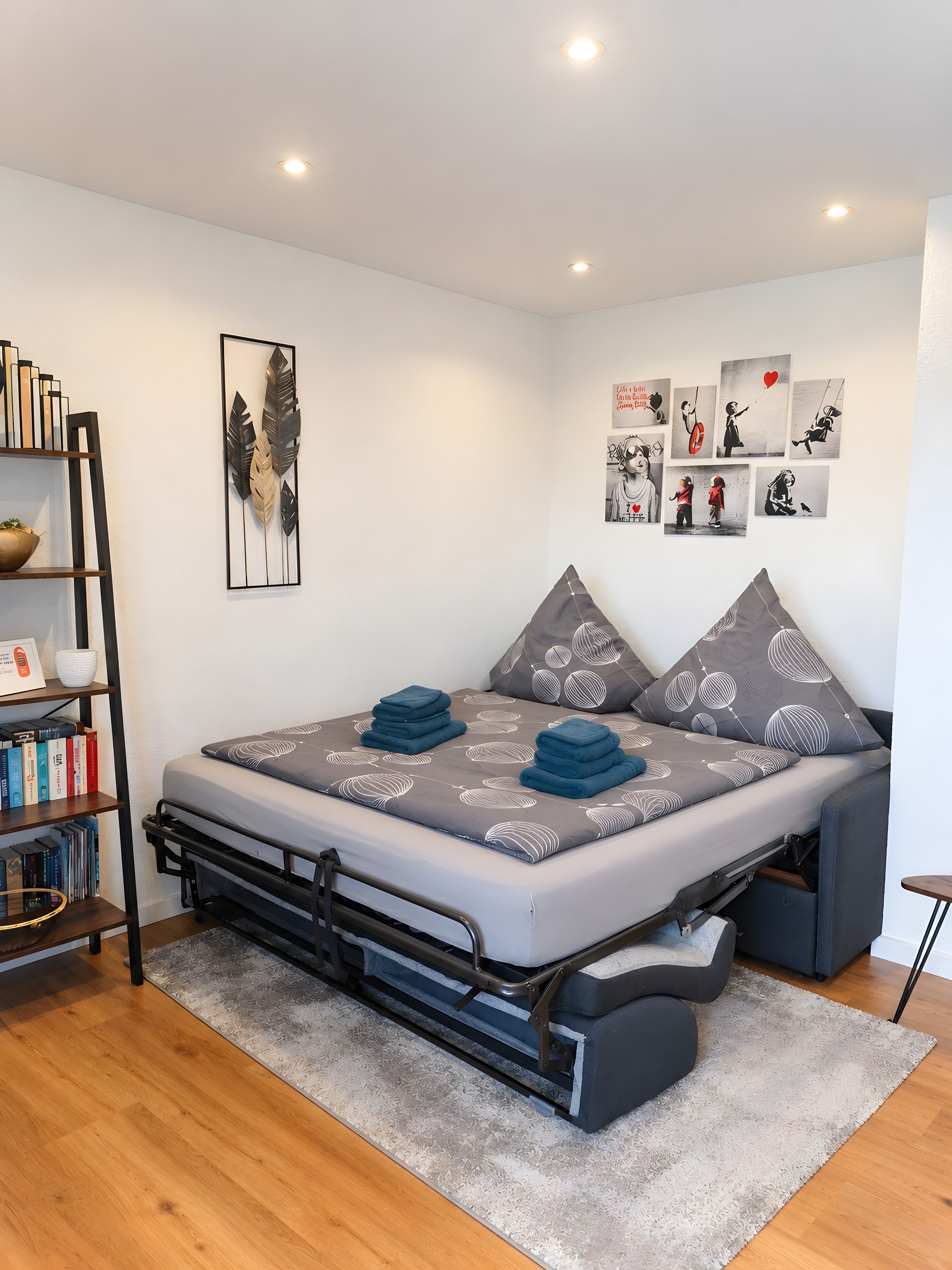 A compact bed with a grey and white leaf-patterned mattress, a small bedside table, and a bookshelf with books and decorative items.