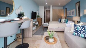 A modern living room with a sofa, coffee table, and a TV. There is a desk with a chair and a mirror. The room has blue walls and a wooden door. - Stylish Studio in Playa del Carmen with Rooftop Pool Walk to Beach & 5th Avenue (Playa del Carmen)