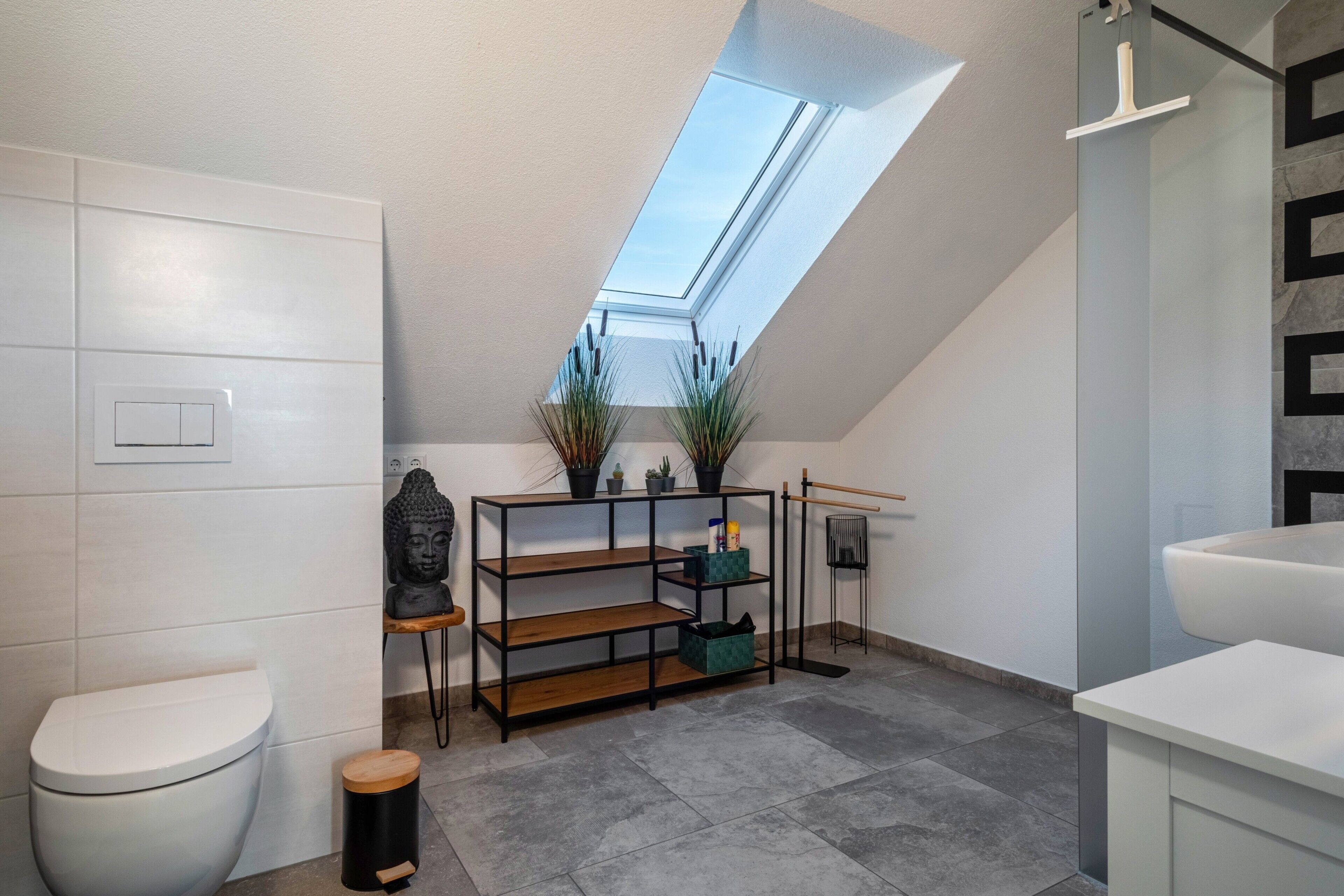 Une salle de bain moderne avec un puits de lumière, des toilettes, une étagère avec des plantes et une baignoire.