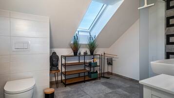 Une salle de bain moderne avec un puits de lumière, des toilettes, une étagère avec des plantes et une baignoire.