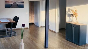 Un salón amplio con suelo de madera, una mesa de comedor con sillas, un alto pilar negro y un cuadro de un muelle.