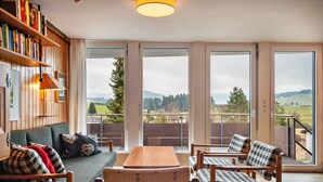 A cozy room with a sofa, chairs, a table, and a bookshelf. Large windows offer a view of the outdoors. - Holiday Apartment "Rommel" with Mountain View, Private Terrace & Wi-Fi (Stiefenhofen)