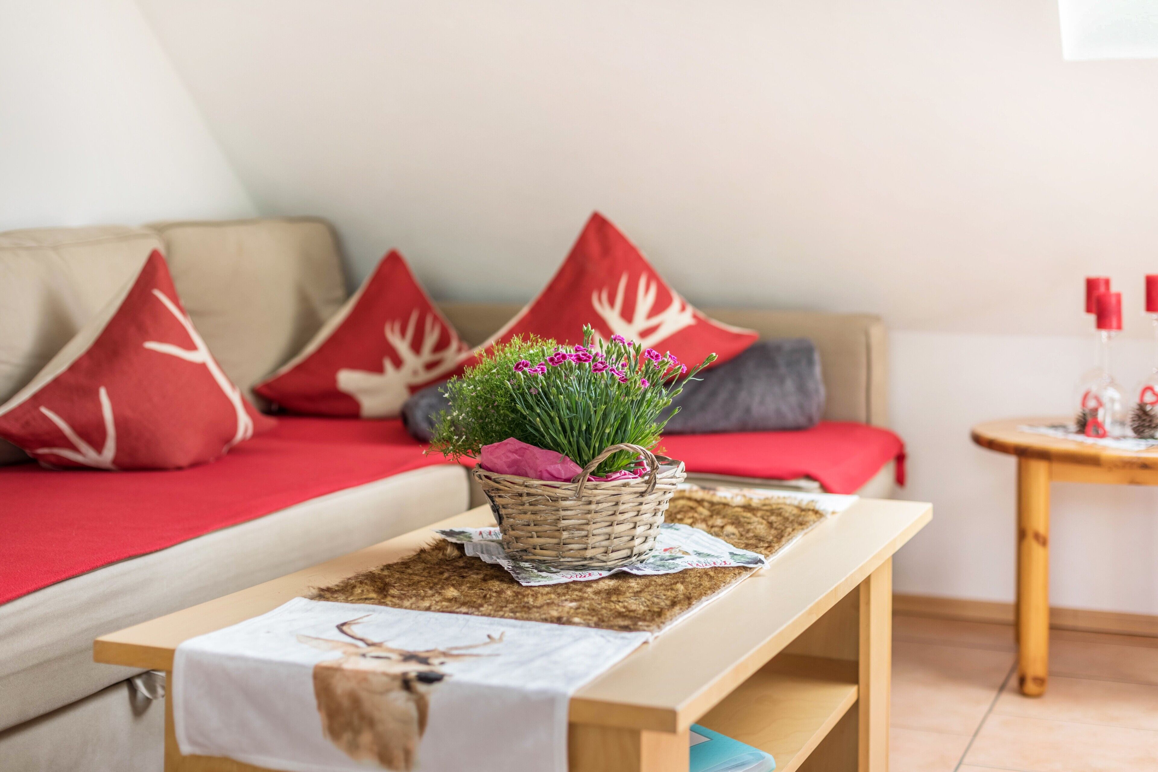 Un salon avec un canapé, des coussins rouges et blancs, une table basse en bois recouverte d’une nappe à motif de cerf, et une petite table avec des bougies rouges.