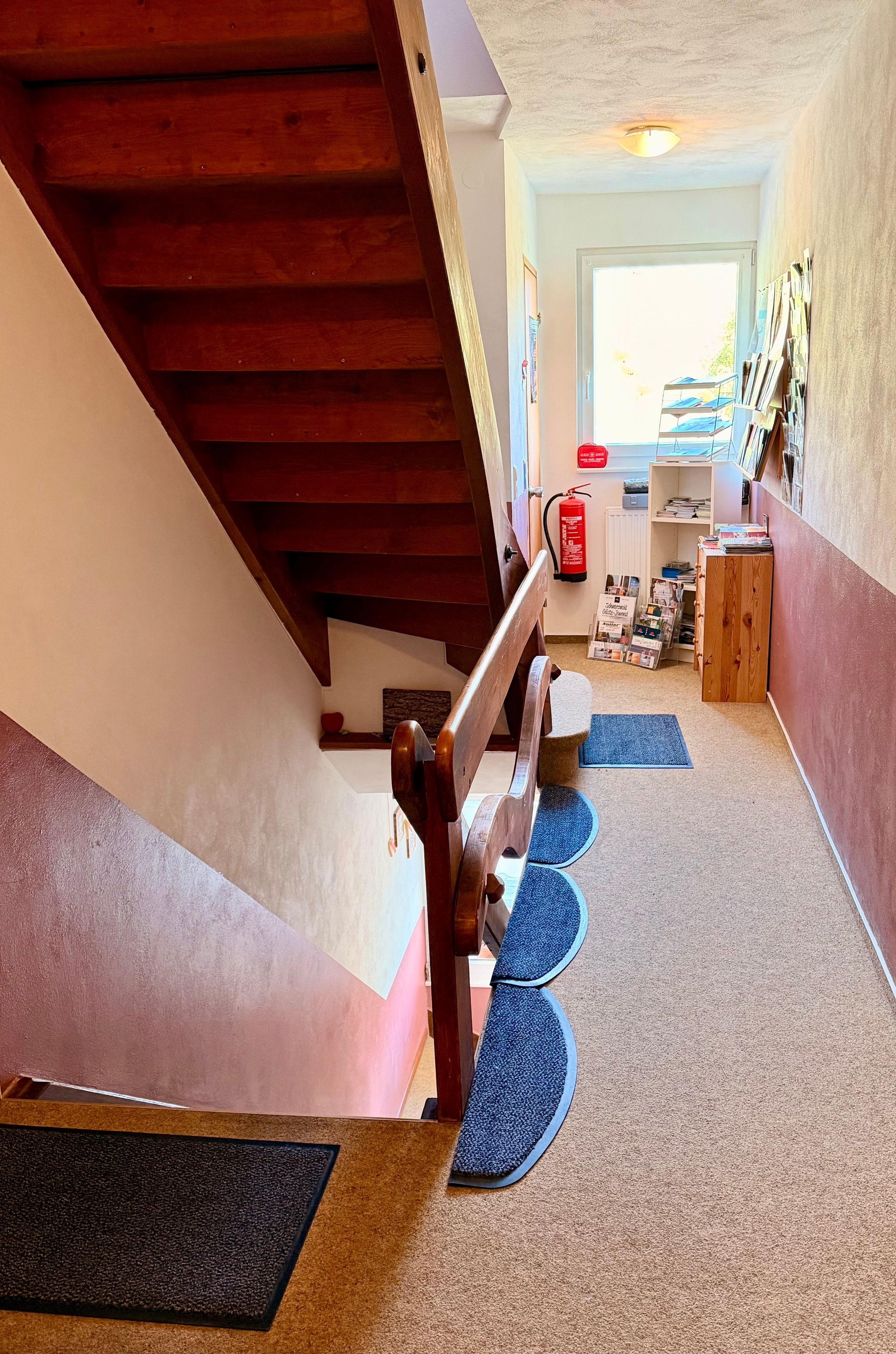 Eine Treppe mit hölzernem Geländer führt zu einer Zwischenebene. Der Boden ist mit blauen Matten ausgelegt, an der Wand ist ein Feuerlöscher angebracht und ein Regal mit Büchern und einem kleinen Tisch vorhanden.