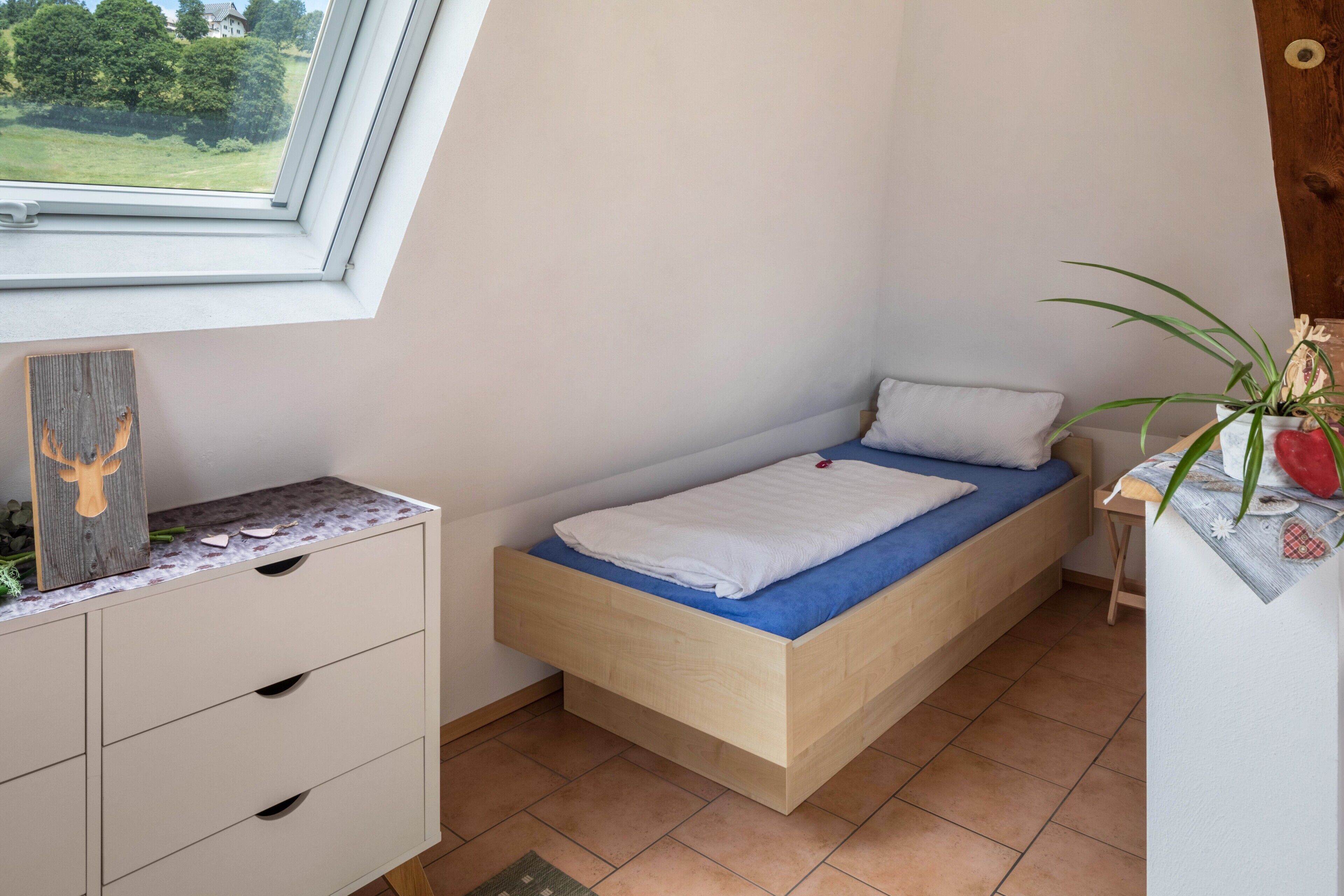 Una camera piccola con un letto singolo, una testiera in legno, una finestra vista verde, un comodino con una pianta e un comò bianco con cassetti.