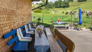 Un patio con panche e un tavolo, un ombrello verde e vista su un prato con delle mucche.