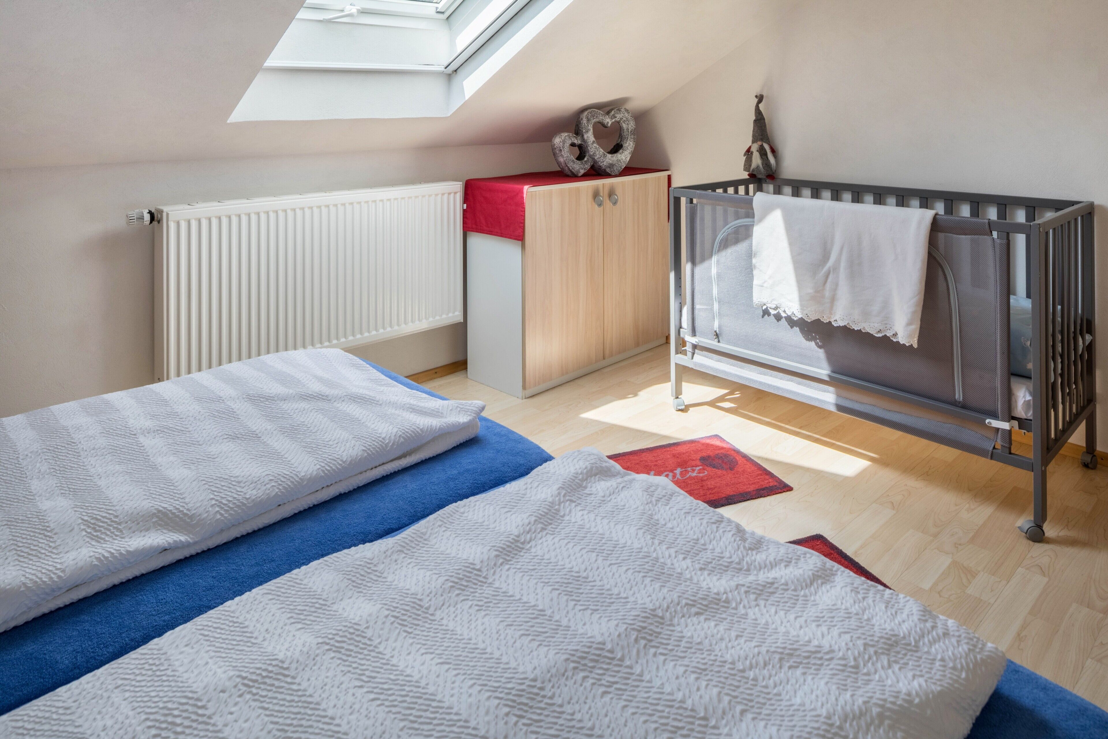 Ein Zimmer mit Oberlicht, einem Babybett, einem Kleiderschrank und einem Bett mit einer blau-weiß gestreiften Decke.