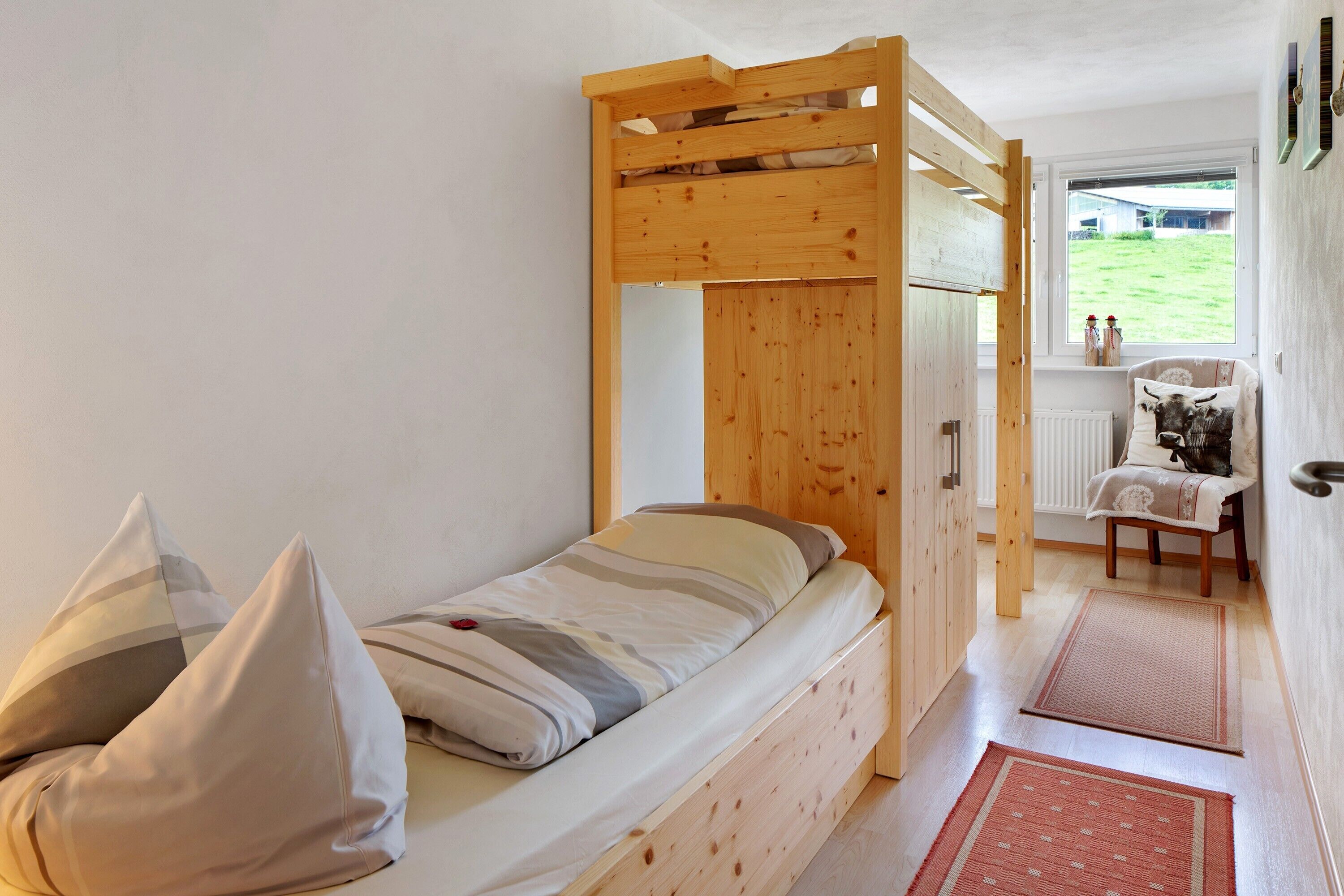 Ein Etagenbett mit Holz-Kopfteil, ein Tisch mit Stuhl und Blick auf einen grünen Garten durch ein Fenster.