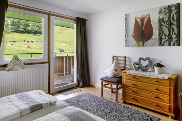 Une chambre à coucher avec un lit, une commode, une chaise, une fenêtre donnant sur un champ où l’on peut voir des vaches, et un tableau.
