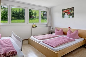 A room with two beds, a window with a view of greenery, a radiator, and a picture on the wall.