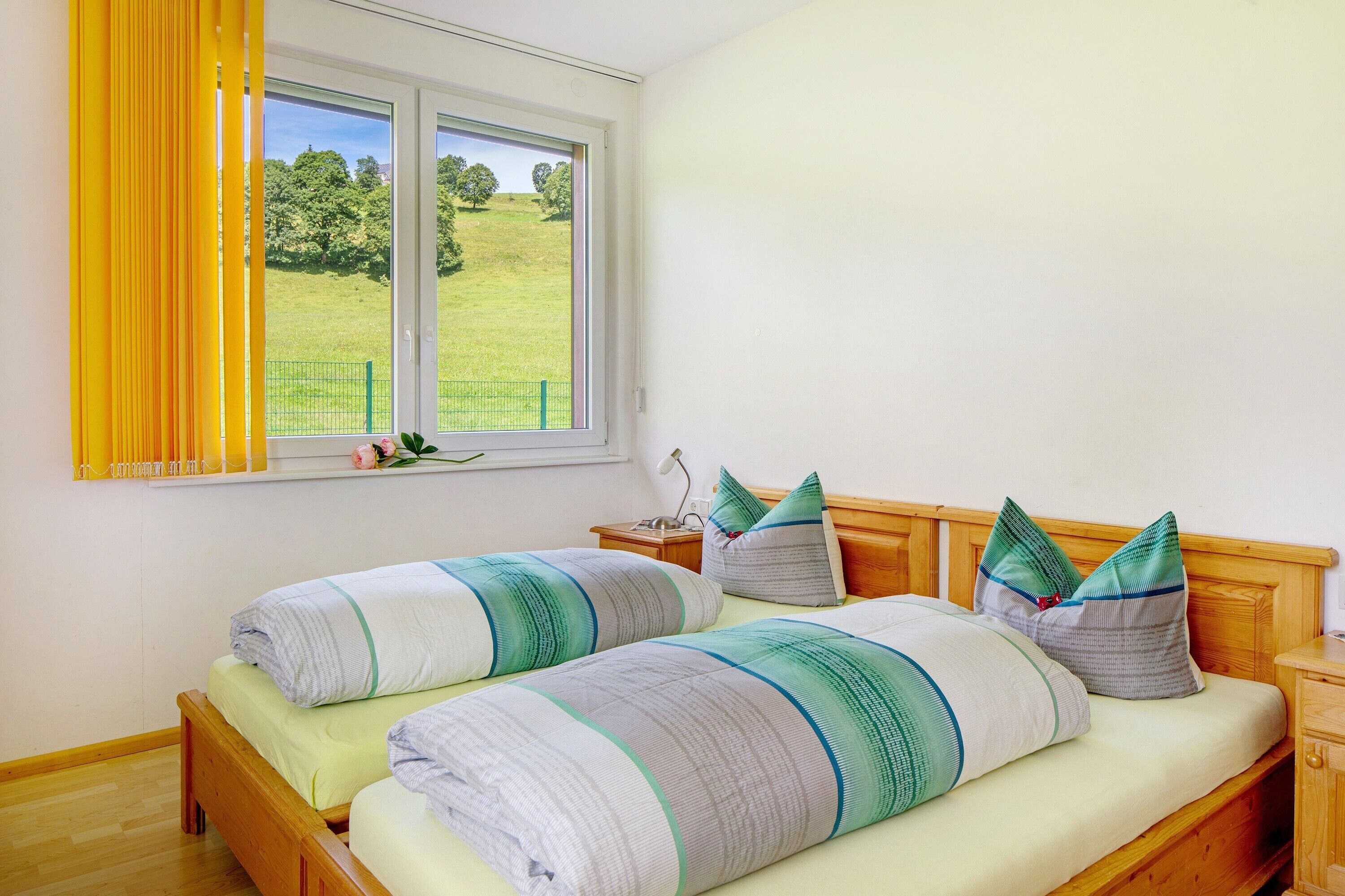 A bedroom with two beds, a window with curtains, a bedside table with a lamp, and a view of a green field.