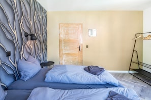 A bedroom with a bed, pillows, a wall-mounted light, a wooden door, and a metal rack with clothes. - Vacation home Haus Waldberg with large garden and covered veranda (Unterschönau)