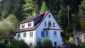 Une maison à deux étages avec un toit rouge, entourée d’arbres et de verdure.