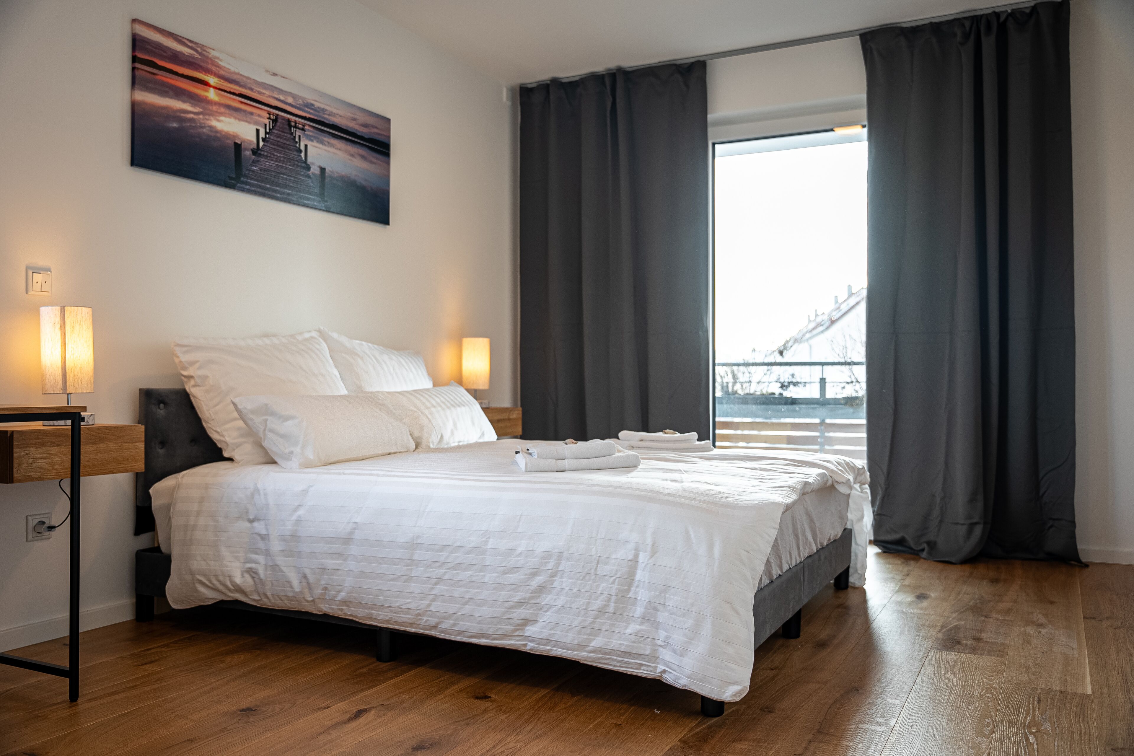 Ein ordentlich bezogenes Bett mit weißen Leinen, ein Nachttisch mit Lampe und ein großes Fenster mit Vorhängen.