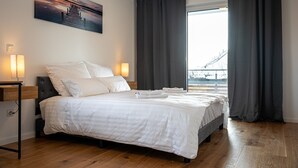 Una cama bien tendida con sábanas blancas, una mesita de noche con lámpara y un amplio ventanal con cortinas.