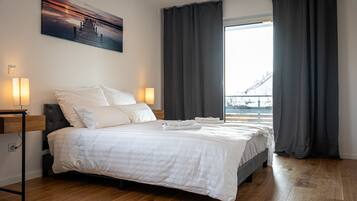 Ein ordentlich bezogenes Bett mit weißen Leinen, ein Nachttisch mit Lampe und ein großes Fenster mit Vorhängen.