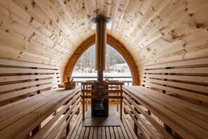 Una cabaña de madera con chimenea central, bancos y un amplio ventanal arqueado.