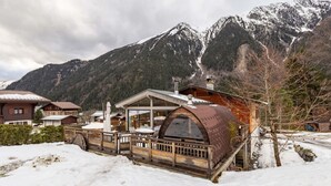 Un edificio de estilo chalé con terraza de madera y un gran ventanal, rodeado de montañas nevadas.