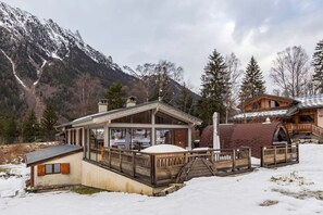 Ein Chalet mit Holzbalkon und Glaseinfriedung, umgeben von schneebedeckten Bergen und Bäumen.