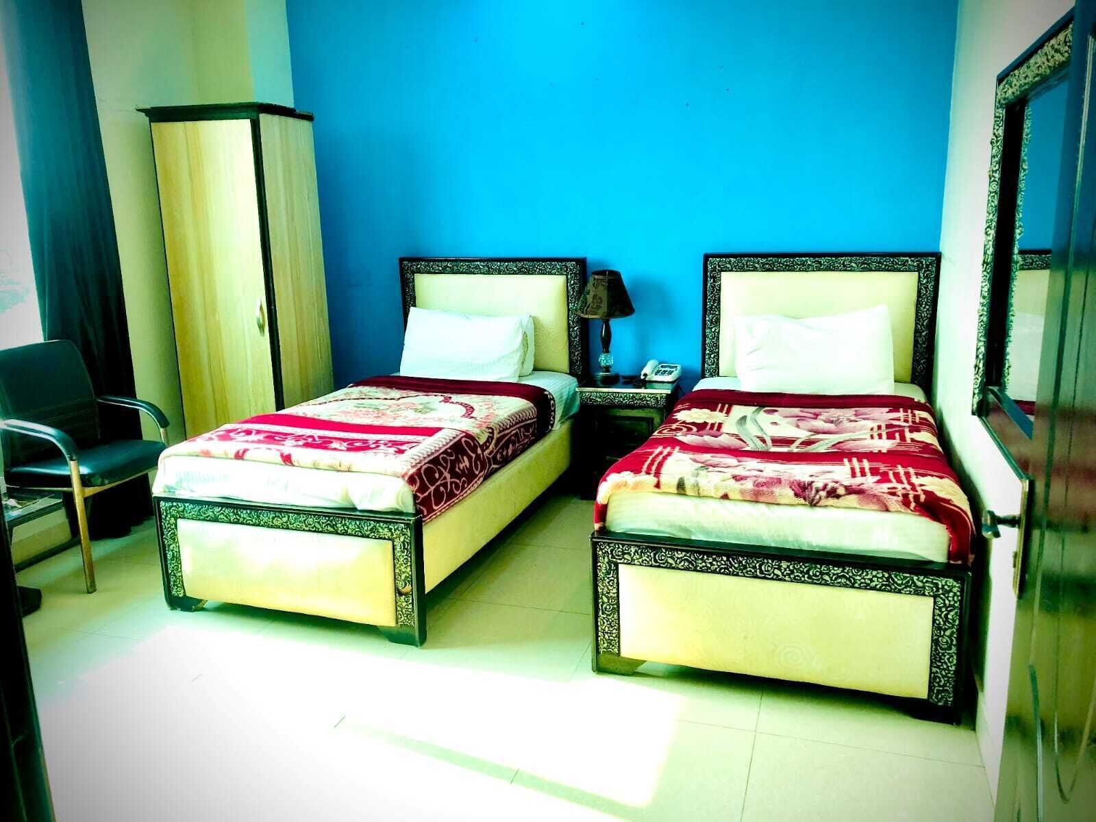 A room with two beds, a chair, a wardrobe, and a nightstand with a lamp.