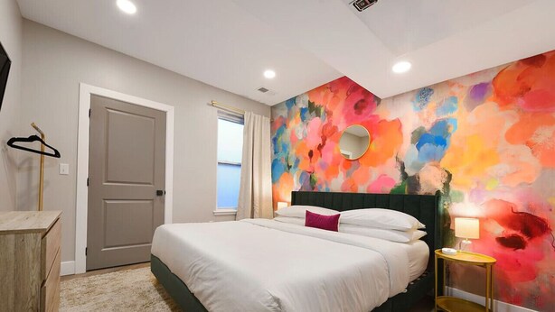 Een slaapkamer met een bed, nachtkastje, spiegel en een kleurrijke abstracte wand.