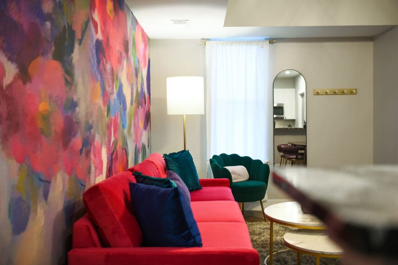 A modern living room with a red sofa, green armchair, and a round coffee table. A colorful abstract painting adorns the wall, and a large mirror reflects the room.