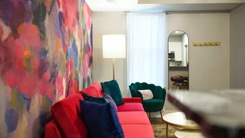 A modern living room with a red sofa, green armchair, and a round coffee table. A colorful abstract painting adorns the wall, and a large mirror reflects the room.