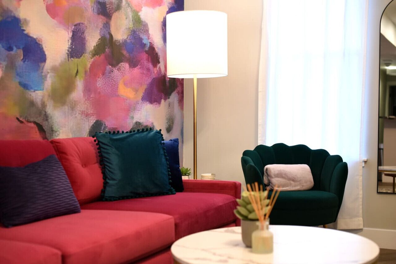A red sofa with teal and blue cushions, a green armchair, a round white table with a plant, and a large abstract painting on the wall.