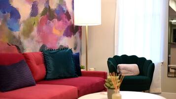 A red sofa with teal and blue cushions, a green armchair, a round white table with a plant, and a large abstract painting on the wall.