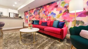 Een moderne woonkamer met een levendig abstract schilderij, een rode bank, een groene fauteuil en een ronde salontafel.