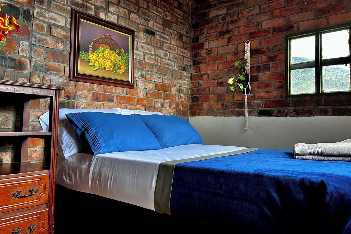 A room with a brick wall, a bed with blue bedding, a wooden dresser, and a framed painting of flowers.