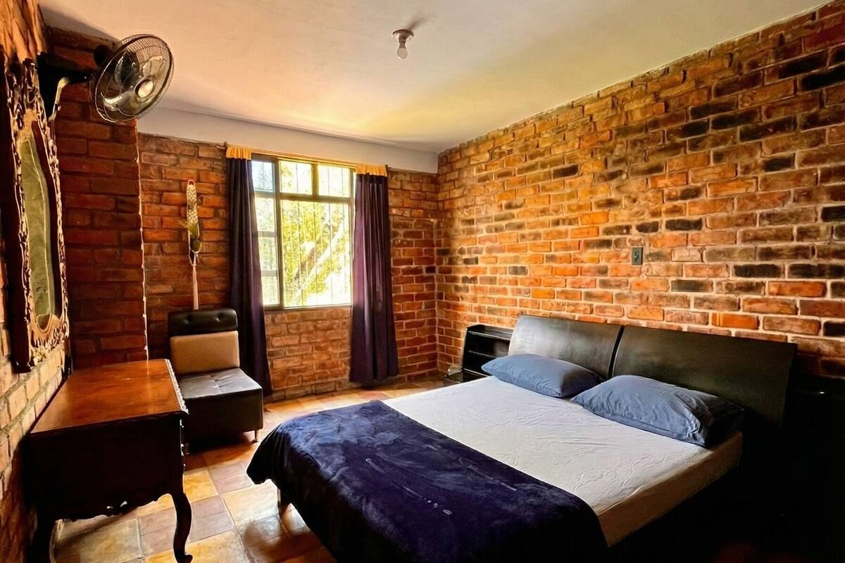 A room with a brick wall, a bed with a headboard, a nightstand, a table, a chair, and a window with curtains.