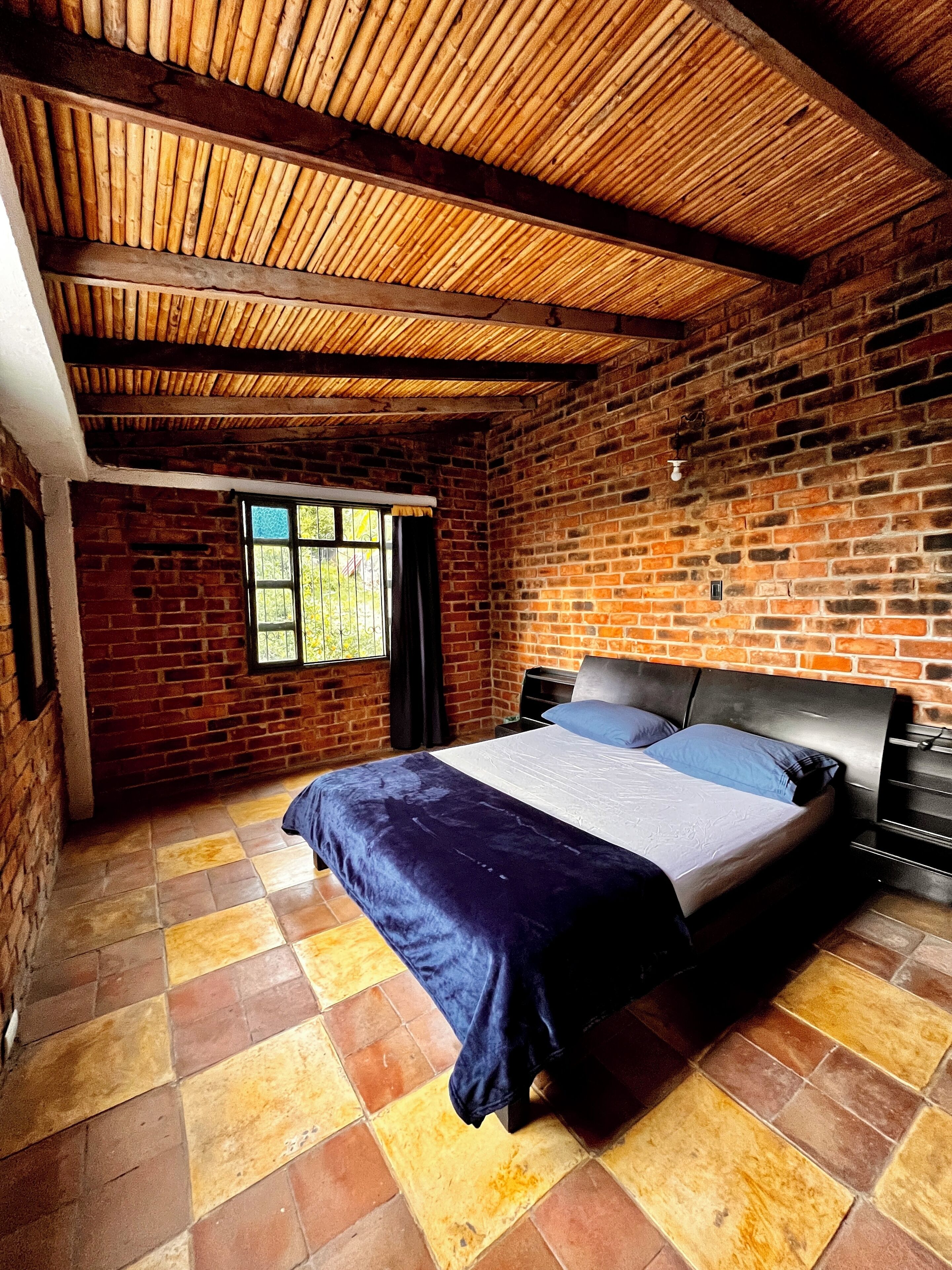 A room with a brick wall, a wooden ceiling, a bed with a headboard, and a window with curtains.