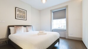 Un lit bien fait, avec une tête de lit, une œuvre d’art abstraite encadrée accrochée au mur, et une fenêtre donnant sur des bâtiments à l’extérieur.