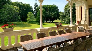 Um pátio externo com mesa de madeira e cadeiras de vime, cercado por colunas brancas e vista para um jardim exuberante.
