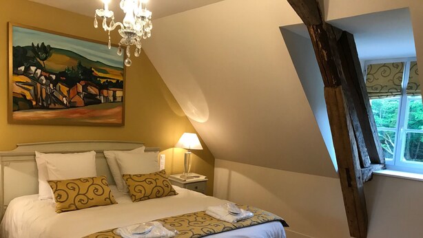 Quarto com cama grande, mesa de cabeceira com abajur, banco com toalhas, quadro na parede e janela com cortinas.